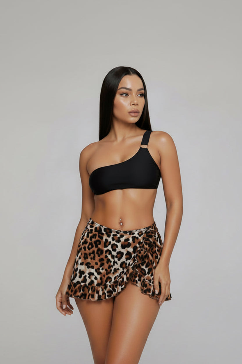 A woman modeling a two-piece swimsuit featuring a black one-shoulder bikini top and high-waisted leopard print mini skirt bikini bottoms, set against a solid gray studio background.