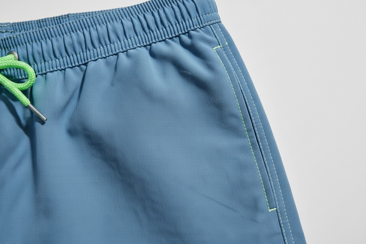 Close-up of slate blue swim shorts showing the elastic waistband, a bright green drawstring, and light green contrast stitching on a light gray background.