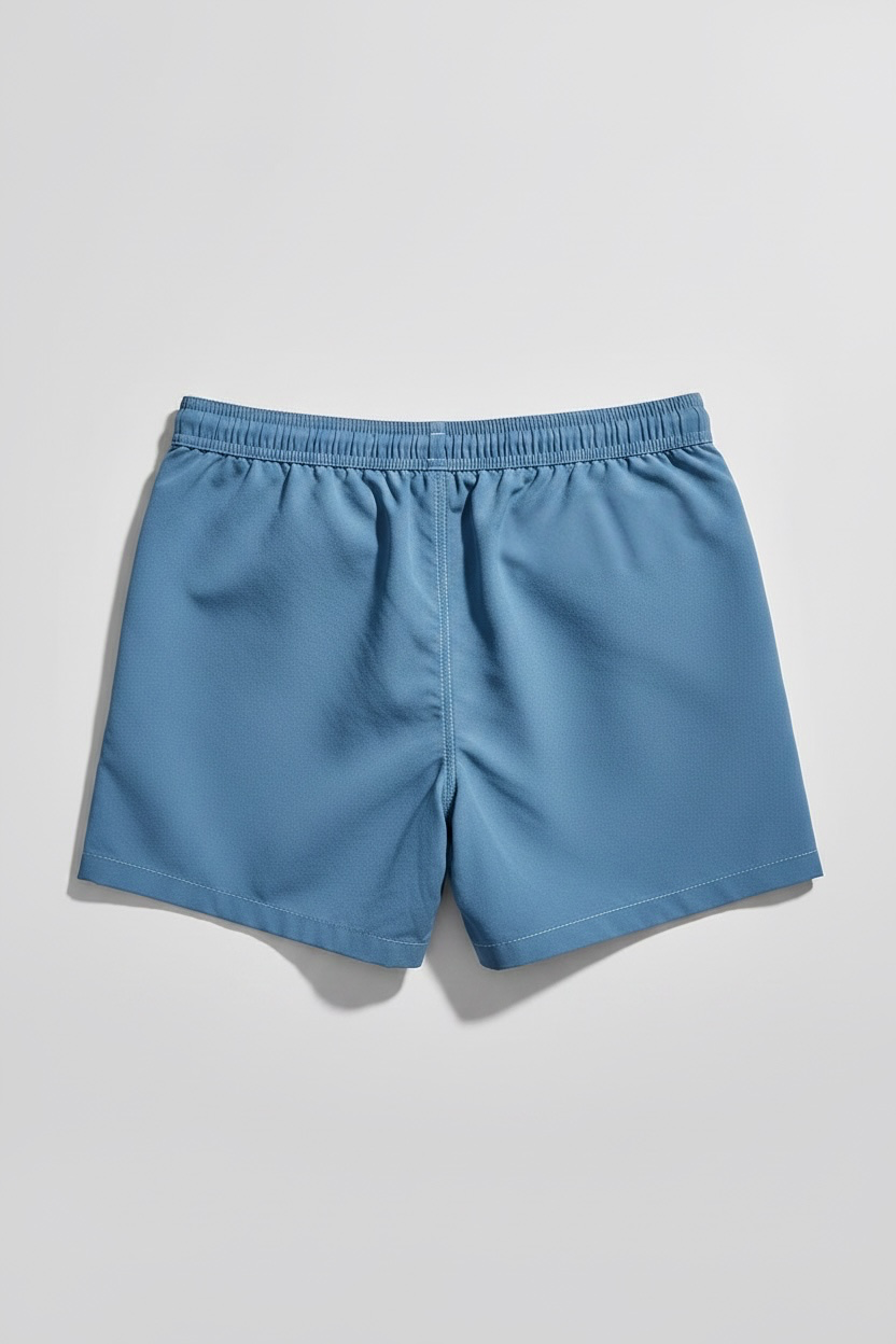 A pair of blue swim shorts with an elastic waistband, laid flat on a light gray background. Back view 