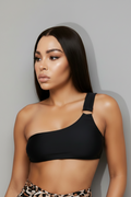 Model wearing a black one-shoulder bikini top paired. The look is completed with rectangular tortoise sunglasses against a light gray studio background.